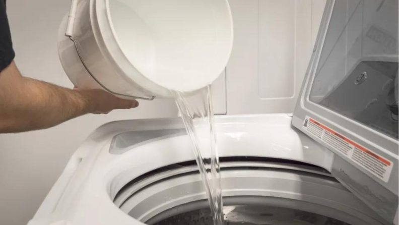 Water from a white bucket being poured into a top loading washing machine