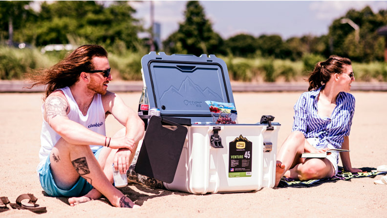 Two people on a beach, sitting beside the Otterbox Venture 45 cooler.
