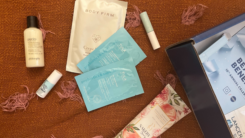 An image of several different makeup and skincare products on an orange bedspread, including a purity skin cleanser, three cleansing masks, a Nakery Beauty rose-scented body and hand wash, and a Tarte Sea mascara tube.