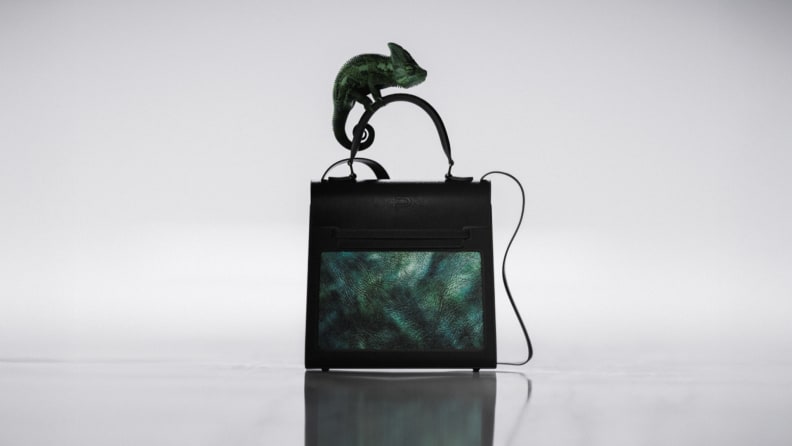 A futuristic black smart handbag features a built-in OLED panel displaying a green pattern, with a small chameleon perched on the handle against a minimalist backdrop.