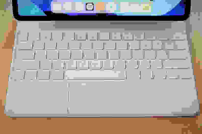 A close-up of Apple's Magic Keyboard (white), showing the layout of keys, as well as the trackpad. The bottom portion of the iPad's display is in frame, but only enough to see iPadOS's dock of apps.