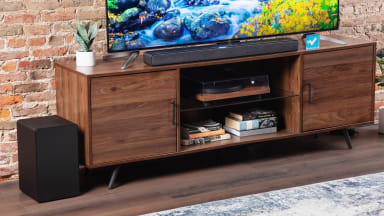 TCL Q Class 3.1 Soundbar sitting on top of wooden credenza in front of flat screen television and acrylic paper weight with blue checkmark in the middle.
