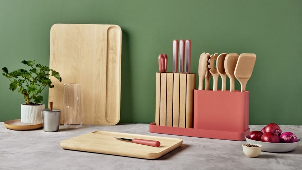 Two wooden cutting boards next to wooden cooking utensils, a knife block, a potted plant, a bowl of onions and a glass vase.