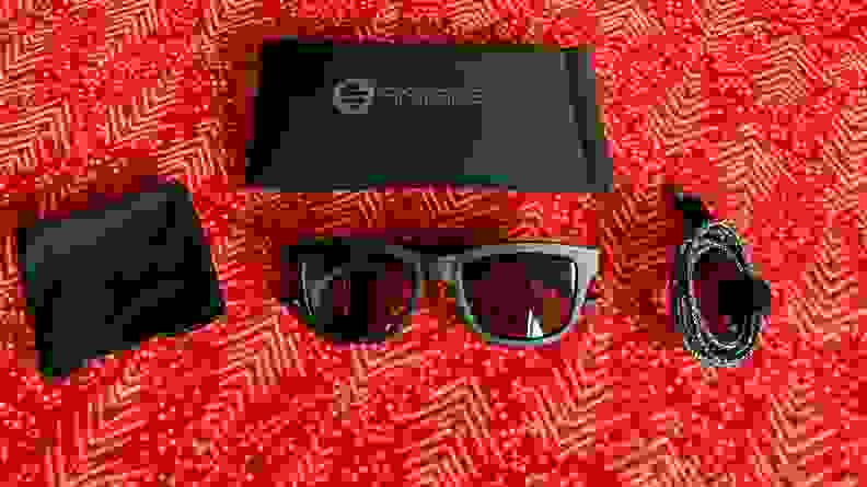 Sunglasses sit on a red background with a shammy, case, and charging cable.
