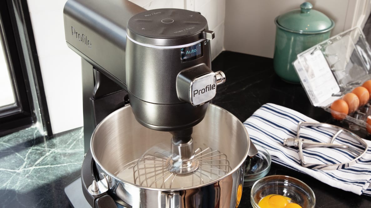 A dark grey GE Smart Stand Mixer sits on a grey counter with the whisk attachment and whipped egg whites inside the mixer bowl.