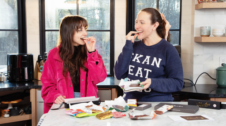 Two people biting into chocolate in front of a counter filled with chocolates