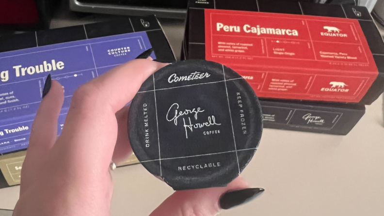 An image of a George Howell Cometeer coffee pod held in a hand over a stack of Cometeer coffee boxes.