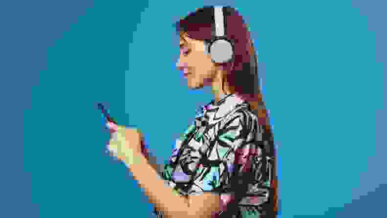 Woman wearing headphones listening to content on her smartphone in front of a blue background.