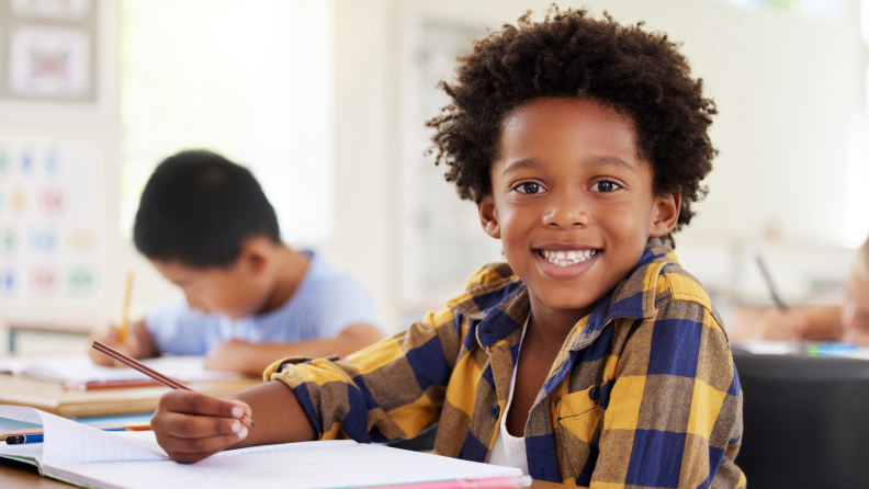 A small child smiling while sitting at a desk in classroom with a pencil and notebook.