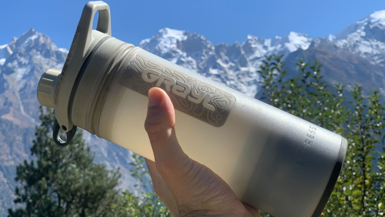A hand holds a white Grayl Geopress filtered water bottle up to the light in front of a view of the Himalayas.