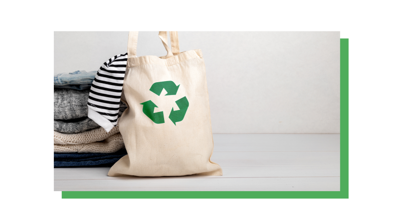 Folded laundry on a white background. A cream laundry hamper with a green recycling logo leans against the pile of clothes and a striped shirt is hanging out of the bag.
