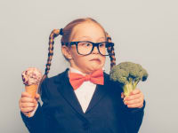 Little girl holds ice cream in one hand, broccoli in the other hand, making a silly face at the broccoli.