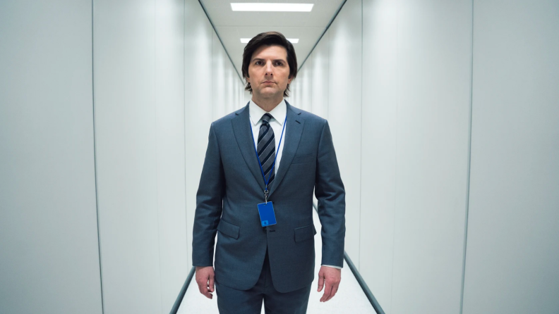 Adam Scott dressed in a suit walking down a white office hallway.