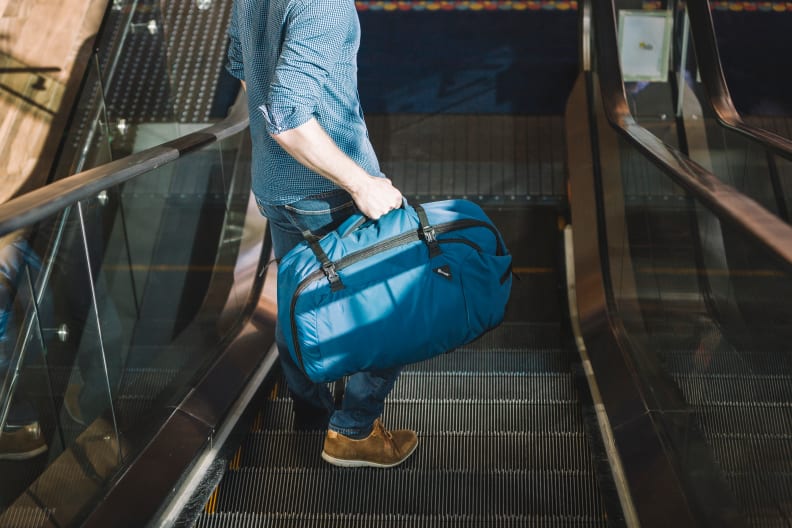 A person carrying a blue PacSafe backpack with his hand down an escalator.