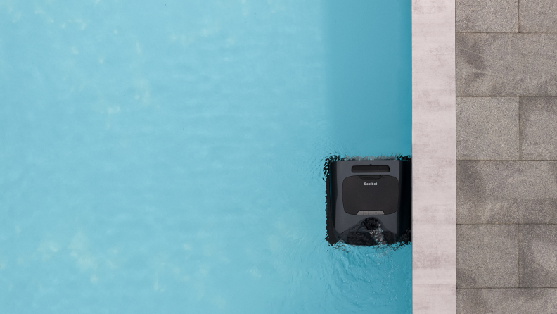 A black, square robot sits on the surface of the water by the edge of a swimming pool