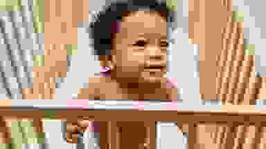 A small child stands up in a wooden crib.