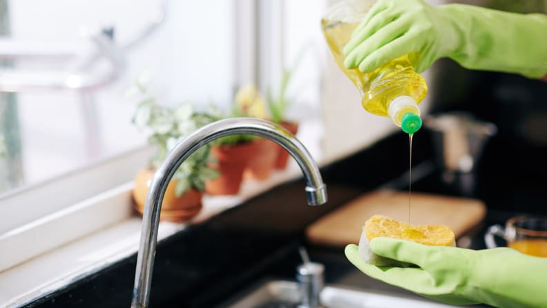 Person wearing green rubber gloves squeezing dish soap onto sponge.