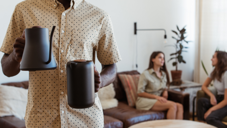 A person is holding a Fellow electric kettle on one hand and a Fellow French press on the other hand. Two people were chatting in the back.