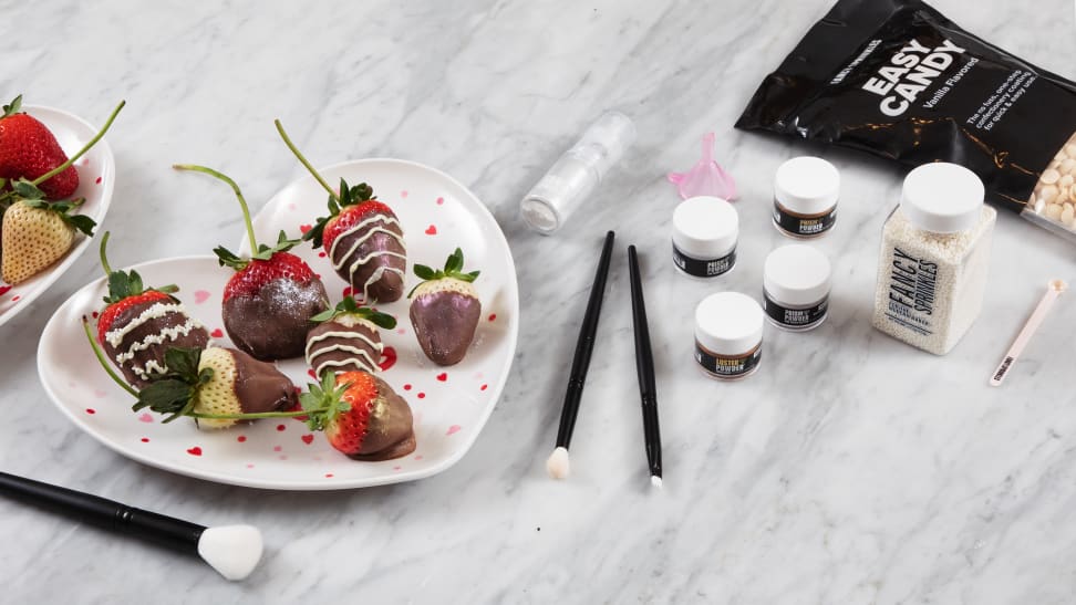 Chocolate covered strawberries on a heart-shaped plate, brushes, pots of edible glitter, and a bag of white chocolate chips sit on a marble counter.