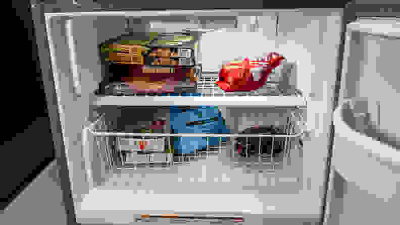 A close-up of the freezer. Inside is a single wire latticework shelf, above a basket  made from the same kind of wire. The drawer and shelf are stocked with frozen foods.