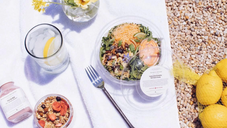 Sakara packaged salad on an outdoor picnic blanket surrounded by  lemon water,  juice, and cereal.