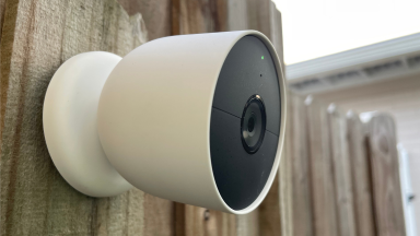 Google's Nest Cam (battery) home security camera hangs on a wood fence outdoors.