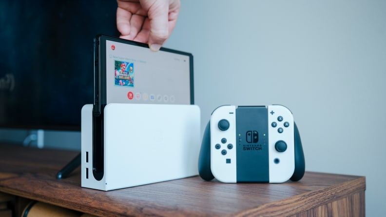 A nintendo switch oled in dock next to a TV with joy-con controller next to it