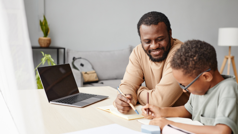 Parent helping child with schoolwork at desk in front of laptop computer.