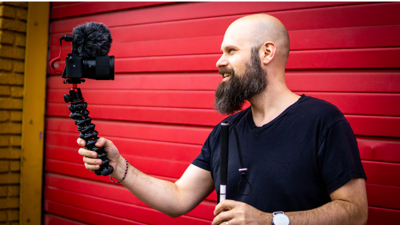 Sam Seavey shown standing in front of a red garage door holding a camera on a stand.