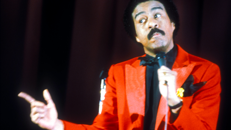 Famed comedian Richard Pryor performs live on stage in a bright-red suit, a finger pointed at the audience.