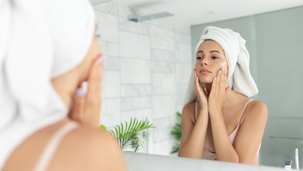A person wearing a white towel on their head and looking into a mirror while touching their cheeks.