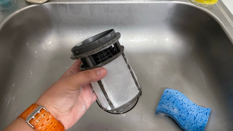 A hand holding a dishwasher filter in the sink with a sponge.