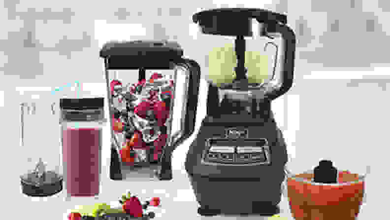 An empty cup. blender, a cup holding a light purple smoothie with a straw, a blender jug holding ice, blueberries, and strawberries, a Ninja blender holding ice cream, and a food processor bowl holding soup on a kitchen counter.