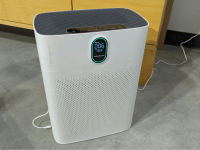 The white air purifier sits on a cement floor, its front face has perforations, welcoming smoke.