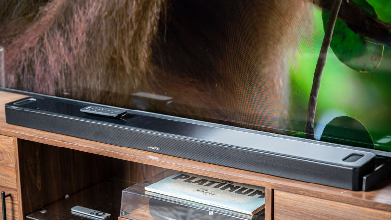 The glass-topped, all-black Bose 900 soundbar sits atop a wooden TV console with media beneath it and a TV behind it