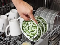 A person loading a bowl covered with spinach puree into a dishwasher that also has plates and mugs in it.