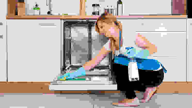 A woman crouches besides a dishwasher with a sponge and cleaning solution bottle. She's scrubbing the interior of the dishwasher's door.