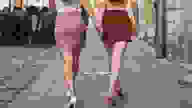Two women walking, facing away from the camera. One wears a patterned pink mini skirt and the other wears a red patterned mini skirt.