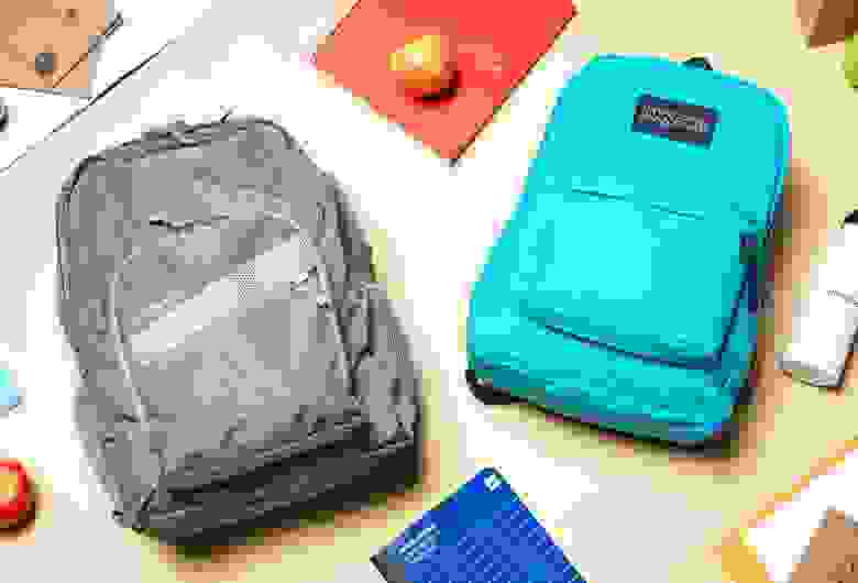 An L.L. Bean backpack and JanSport backpack laying on a table surrounded by school supplies and apples.