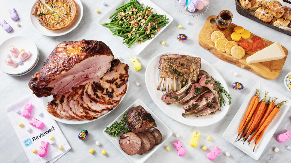 A buffet of Easter dinner options spread on a marble counter: ham, lamb roast, lamb chops, quiche, green beans, carrots, a cheese board, and hot cross buns, surrounded by bits of colorful Easter candy.