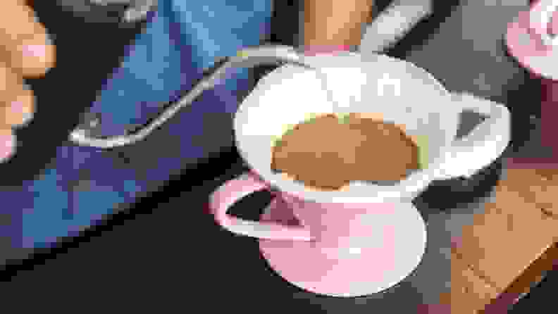 A person pours hot water into a pink pour-over coffee maker lined with a coffee filter and filled with coffee grounds.