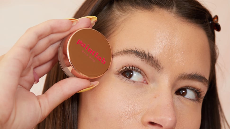 A closeup on the top half of a model's face as she holds a jar of brow soap up to her eyebrow.