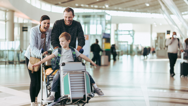 Family of three walking through airport together smiling while son sits on top of suitcase cart.