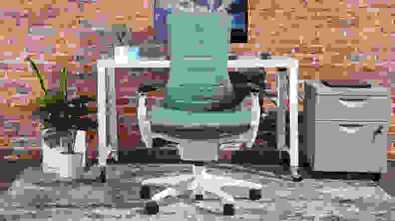 The Herman Miller x Logitech G Embody gaming chair in an office space, next to a white desk, computer monitor, and office supplies.