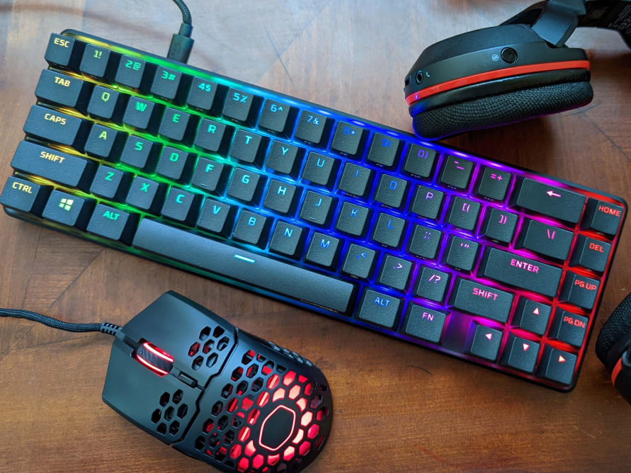 A black keyboard with glowing rainbow colors flanked by a black gaming headset and a black gaming mouse that's glowing red