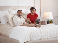 A smiling couple relaxes on a neatly made bed with cream-colored bedding, as one person holds a remote and the other uses a tablet. A wooden nightstand with a lamp and potted plant sits beside the bed in a bright, cozy bedroom.