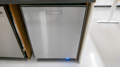 A stainless-steel dishwasher is installed in a cabinet in a lab environment, and its bottom light shines blue.