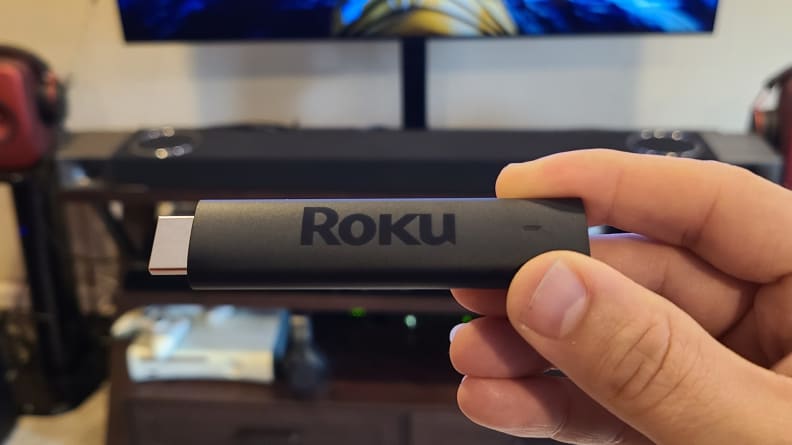 The matte black, candy-bar sized Roku Streaming stick is held with its port showing an OLED TV.