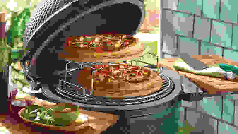 An open Big Green Egg with two pizzas cooking inside, on a tiered grate.
