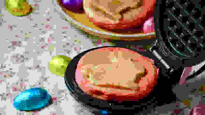 A round snack-sized waffle is in a Dash Bunny mini waffle maker. Some more are in the background, along with Easter chocolates.